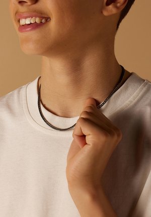 Zilverkleurige kettinghalsband, gelaagd over een witte crewneck t-shirt. De ketting heeft een gladde, gestructureerde uitstraling en past nauw om de hals.
