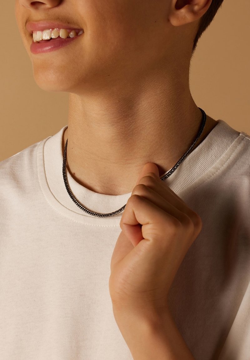 Zilverkleurige kettinghalsband, gelaagd over een witte crewneck t-shirt. De ketting heeft een gladde, gestructureerde uitstraling en past nauw om de hals.