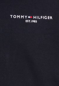 Logo de TOMMY HILFIGER con la bandera roja, blanca y azul y el texto EST. 1985 sobre tela negra