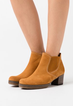 Bottines en suédine beige avec un talon épais, des panneaux latéraux élastiques et des coutures blanches contrastantes. Texture lisse et design à bout arrondi.
