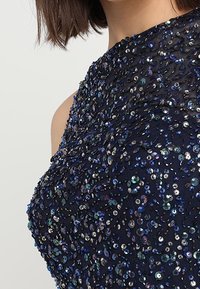 Robe de marine avec des embellissements en perles complexes et des sequins irisés sur un tissu texturé, stylisée avec un design asymétrique.