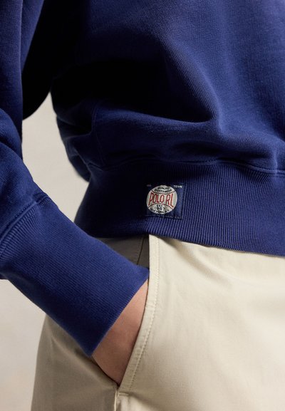 Άτομο με μπλε σκούφο φούτερ με λογότυπο Polo RL και μπεζ παντελόνια, το χέρι μερικώς στην τσέπη.