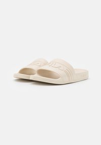 Infradito beige con una larga fascia texturizzata che presenta il logo del marchio in rilievo. Suola piatta con motivo sottile e design open-toe.
