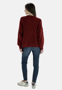 Bordeauxroter Strickpullover mit lässiger Passform, gerippter Textur und überschnittenen Schultern, kombiniert mit dunkelblauslim Jeanz und weißen Sneakers.