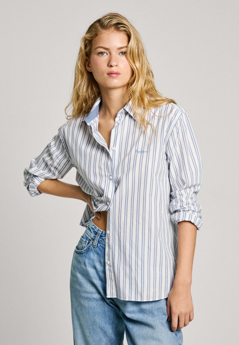Chemise à rayures, blanche avec des rayures verticales bleues. Comprend un col, des manches retroussées et une coupe légèrement oversize. Jean en denim léger.