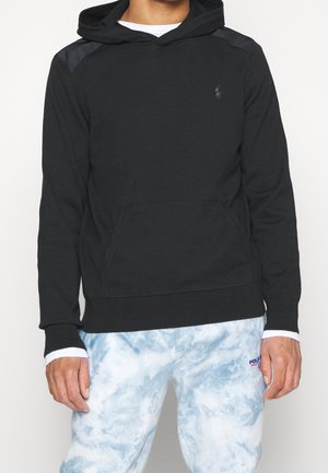 Mężczyzna w czarnej bluzie z kapturem z kieszenią z przodu oraz biało-niebieskich spodniach tie-dye z logo „Polo Ralph Lauren” obok kieszeni.