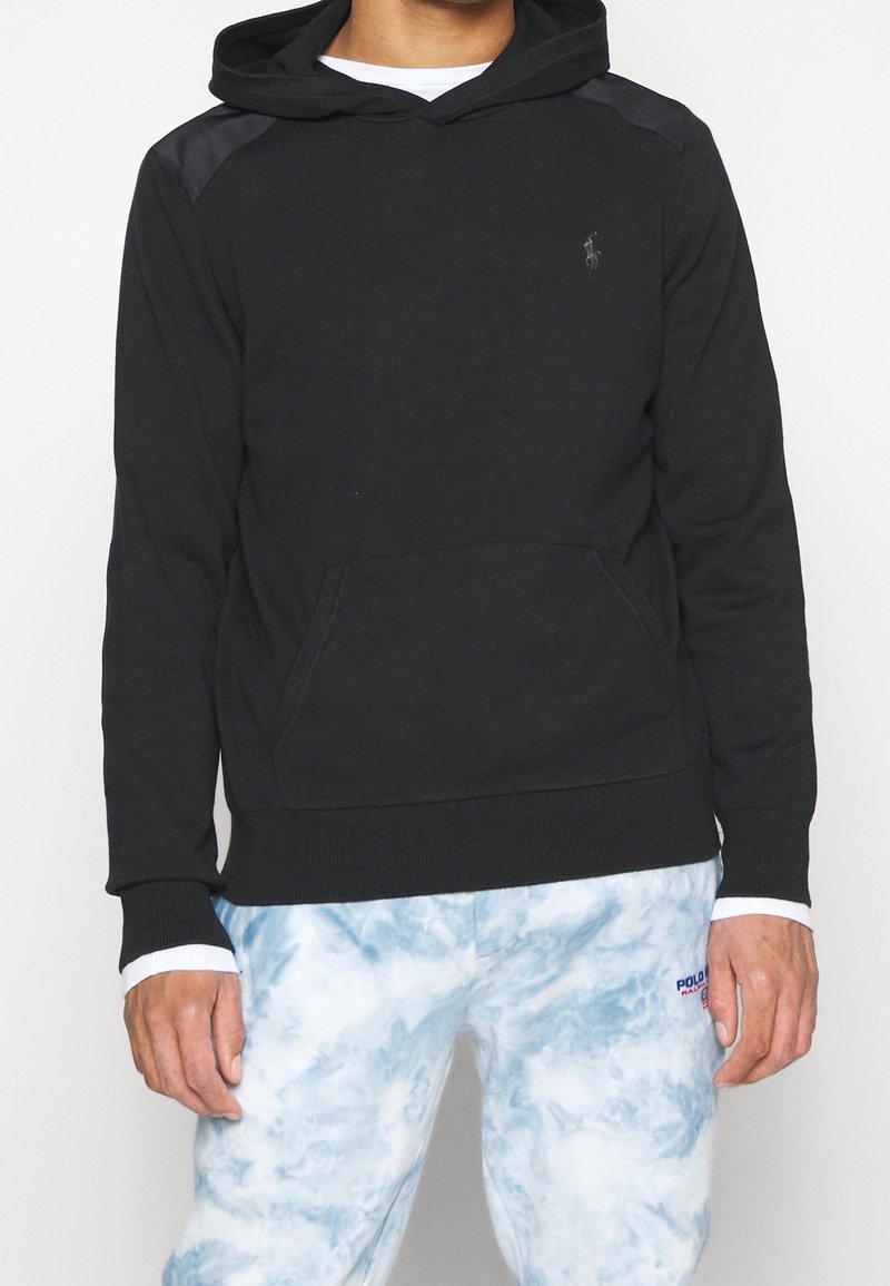 Homme portant un sweat à capuche noir avec poche avant et un pantalon tie-dye blanc et bleu avec le logo "Polo Ralph Lauren" près de la poche.