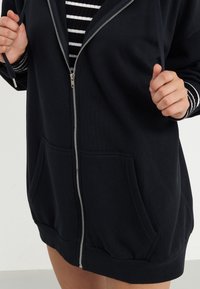 Crna zip-up majica od mješavine pamuka, s prednjim džepovima, rebrastim detaljima i opuštenim krojem s kapuljačom i zatvaranjem na zip.