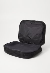 Svart stoff reiseveske med glidelåsåpning, med et mesh-interiørpanel og en flat, rektangulær form for oppbevaring.