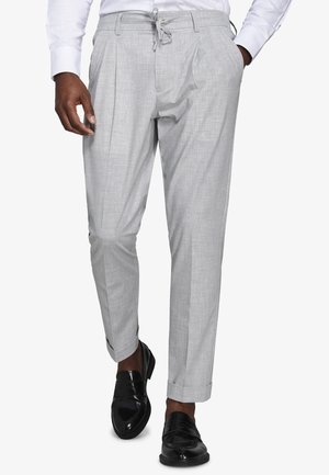 Mężczyzna w jasno szarych, dopasowanych spodniach ze zwiniętymi mankietami, białej koszuli, czarnych skórzanych mokasynach, z jedną ręką w kieszeni, idący do przodu.