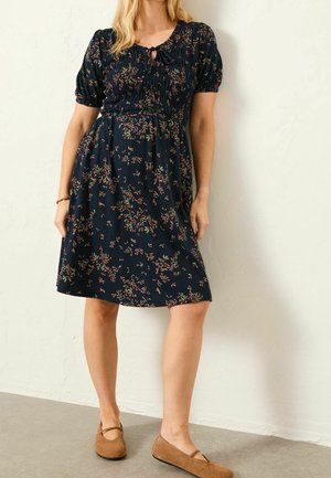 Femme portant une robe bleu marine au genou avec de petits motifs floraux et des manches courtes bouffantes, associée à des chaussures plates marron.
