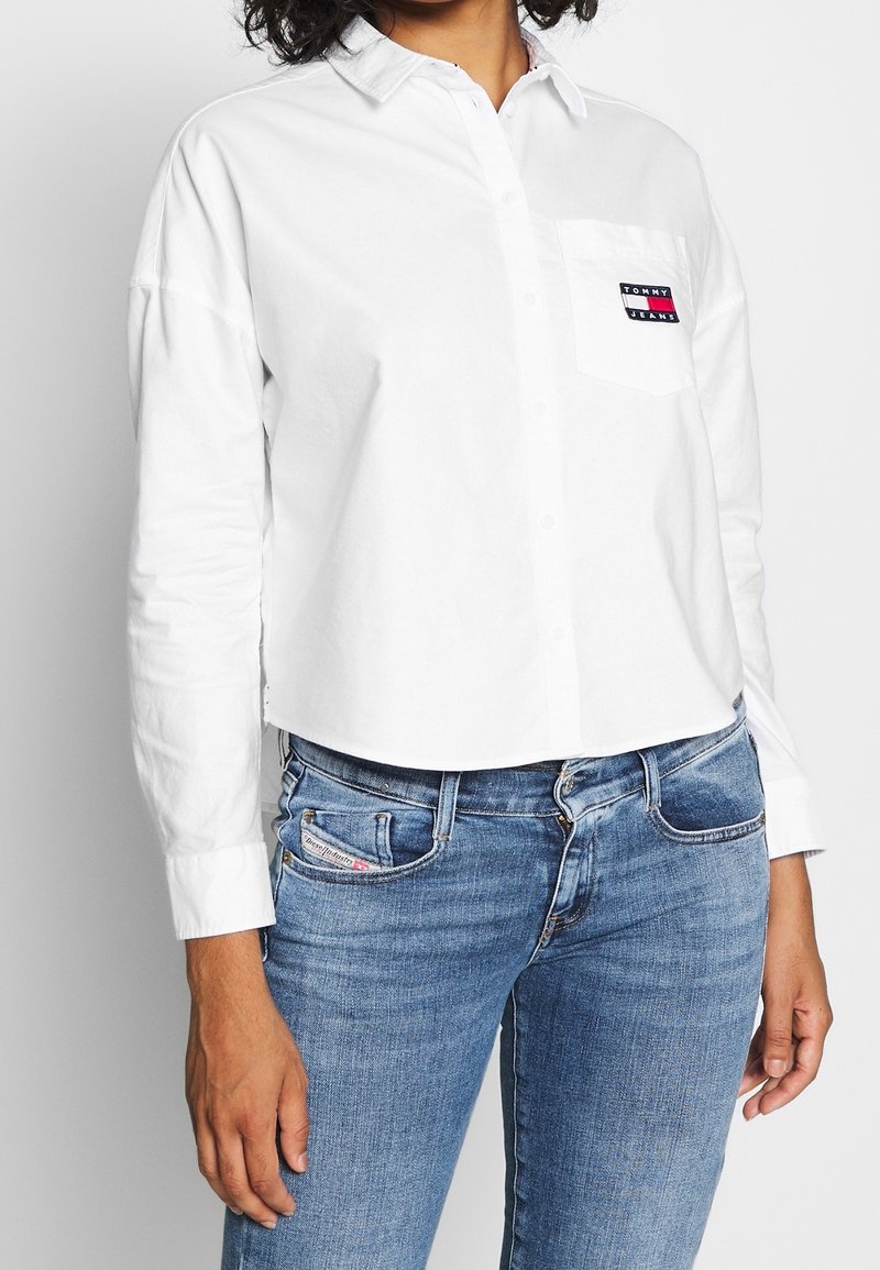 Vit kortärmad skjorta med långa ärmar, knäppning vid kragen och vänster bröstficka med en färgad logotypapplikation. Bärs tillsammans med ljusblå jeans.