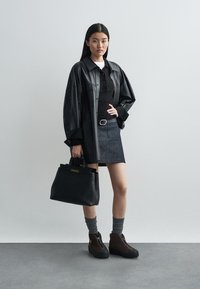 Sort læder oversized skjorte, sort polo, denim nederdel, høje brune støvler, grå strømper, holder en sort håndtaske med guld detaljer.