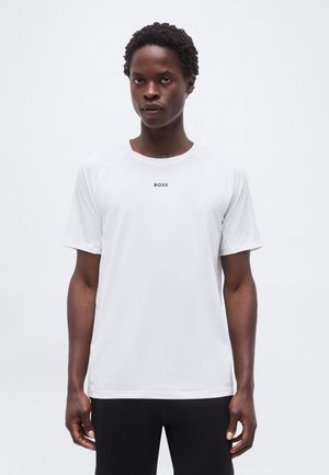 Jeune homme portant un t-shirt de sport blanc à manches courtes avec le logo "BOSS", debout devant un fond clair uni.
