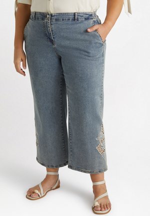 Jeans cropped in denim blu con dettagli floreali intagliati near the hem, dotati di tasche frontali e una chiusura a bottone classica. Abbinati a sandali piatti.