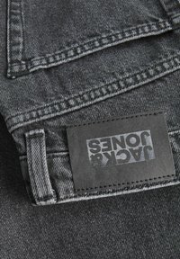Tela de denim gris oscuro con costuras cosidas, presentando un parche de cuero negro etiquetado "JACK JONES". Textura suave, diseño minimalista.
