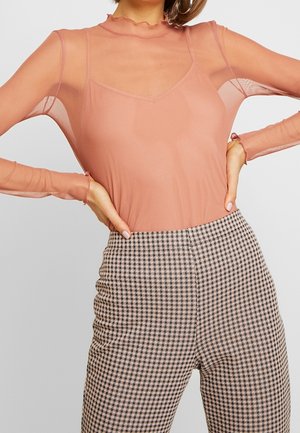 Femme portant une blouse pêche transparente à manches longues avec des poignets à volants sur un caraco pêche, associée à un pantalon taille haute à motif pied-de-poule.