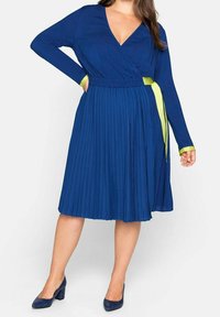 Robe portefeuille plissée bleue à manches longues, avec un décolleté en V profond et une ceinture nouée jaune-vert contrastante à la taille. Texture de tissu lisse.