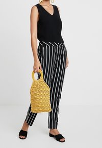Czarny top bez rękawów, czarno-białe spodnie w paski oraz żółta pleciona torebka z okrągłym uchwytem; czarne buty z odkrytymi palcami.