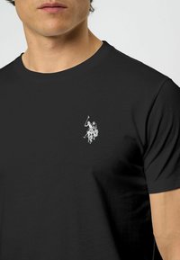 Vīrietis valkā melnu t-kreklu ar mazu baltu polo spēlētāja logo, izšūtu kreisajā krūtī.