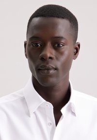 Camicia bianca texturizzata con colletto classico, dotata di abbottonatura e dettagli di cucitura sottili, indossata da un modello con capelli corti.