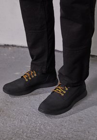 Zapatos negros con una textura suave, con cordones y ojetes naranjas. Llevados con pantalones negros, mostrados desde el tobillo hacia abajo sobre una superficie de concreto.