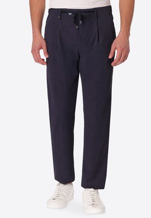 Uomo che indossa pantaloni blu scuro con coulisse in vita e sneakers bianche, in piedi davanti a uno sfondo chiaro e semplice.