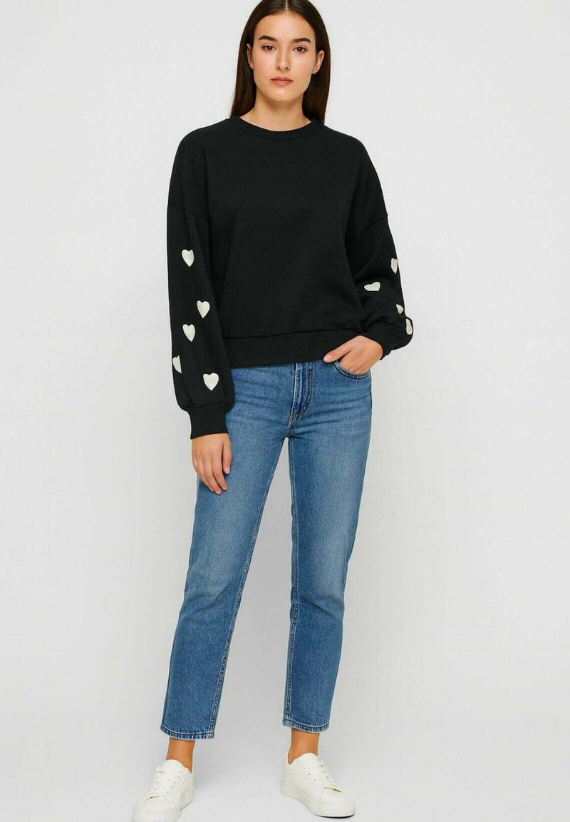 Vrouw in een zwart sweatshirt met witte hartpatronen op de mouwen, blauwe jeans en witte sneakers, staande tegen een effen achtergrond.