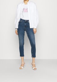 Camisa blanca de botones, camiseta gráfica rosa, jeans de mezclilla cortos azules y sandalias de tacón blancas. Diseño sencillo con un aspecto casual.