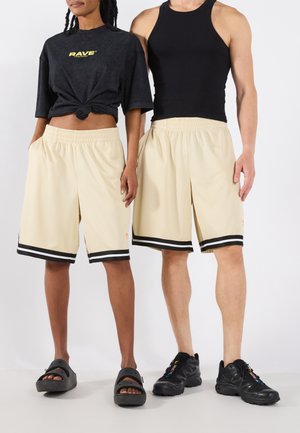 Beige knielange Shorts mit schwarzen Streifen, aus atmungsaktivem Material, kombiniert mit einem schwarzen cropped T-Shirt und einem schwarzen Tanktop.