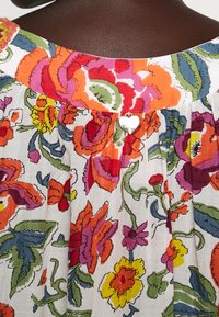 Květinové šaty s bílým pozadím a živými vzory v červené, oranžové, růžové a zelené barvě; doplněné stříbrným detailem ve tvaru srdce na knoflíku.