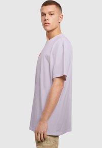 Lilla bomulds-t-shirt med korte ærmer, afslappet pasform, rund halsudskæring, med et rødt logo på brystet. Glat tekstur, minimalistisk design.