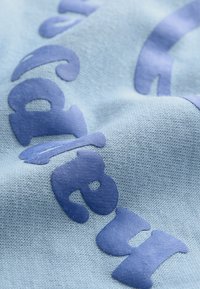 Világoskék, puha tapintású textília, vastag sötétkék betűkkel és játékos formákkal díszítve, amelyek térhatású hatást keltenek.