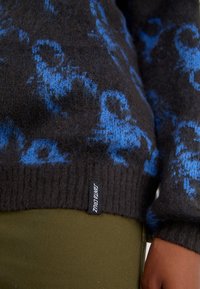 Maglione lavorato a maglia di colore grigio scuro con motivi astratti blu. Polsini e fondo a costine. Etichetta visibile: "Santa Cruz." Abbinato a tessuto verde oliva.