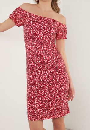 Kobieta w czerwonej sukience z odkrytymi ramionami, krótkimi bufkowymi rękawami i białym kwiatowym wzorem, stojąca na tle jednolitego jasnego tła.