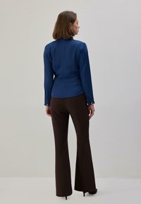 Blusa azul de manga larga con silueta ajustada, puños abotonados, combinada con pantalones marrones acampanados y tacones negros, vista desde atrás.