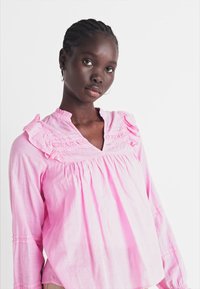 Blusa di cotone rosa con colletto arricciato e yoke frontale. Maniche lunghe con dettagli arricciati. Tessuto leggero, vestibilità comoda.