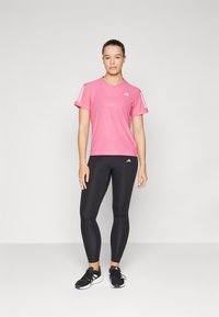 Maglietta sportiva rosa con righe e logo bianche, abbinata a leggings neri. Modello che indossa scarpe da ginnastica nere. Design semplice e aderente.