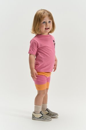 Mali dječak svijetle kose stoji gledajući blago prema gore, nosi ružičastu majicu, narančaste i ružičaste kratke hlače, bež čarape i crno-bijele klizne tenisice sa šahovnicom.