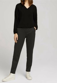 Blusa nera a v con maniche lunghe, abbinata a pantaloni slim-fit grigio scuro. Il modello indossa sneakers alte bianche. Design e colori minimalisti.