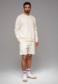 Ensemble en crème comprenant un sweat-shirt et un short en matériau doux. Présente une coupe décontractée, des poignets côtelés et un logo accentué sur le sweat-shirt. Baskets blanches et chaussettes rayées.