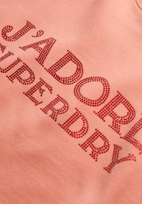 Sudadera rosa con "J'ADORE" y "SUPERDRY" en strass rojos contorneados, con una textura de algodón suave y escote redondeado.