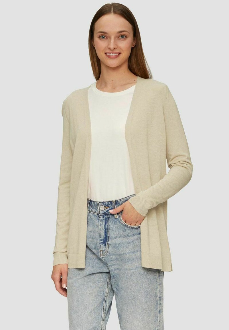 Beige cardigan met open voorkant, lange mouwen en geribde manchetten, gedragen over een wit shirt en gecombineerd met lichtblauwe jeans. Zachte textuur.