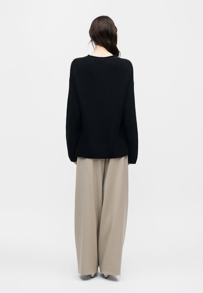 Γυναίκα που στέκεται πλάτη, φοράει μαύρο πλεκτό πουλόβερ και φαρδιά μπεζ παντελόνια σε απλό φωτεινό φόντο.