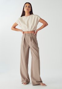 Top corto beige con orlo arricciato, abbinato a pantaloni larghi marrone chiaro. La modella indossa calzature minimal, con le mani sui fianchi.