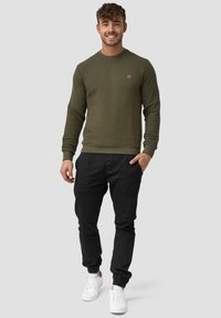 Sudadera texturizada de color verde oliva con cuello redondo, puños y dobladillo acanalados; combinada con pantalones negros tipo tapered y zapatillas blancas.