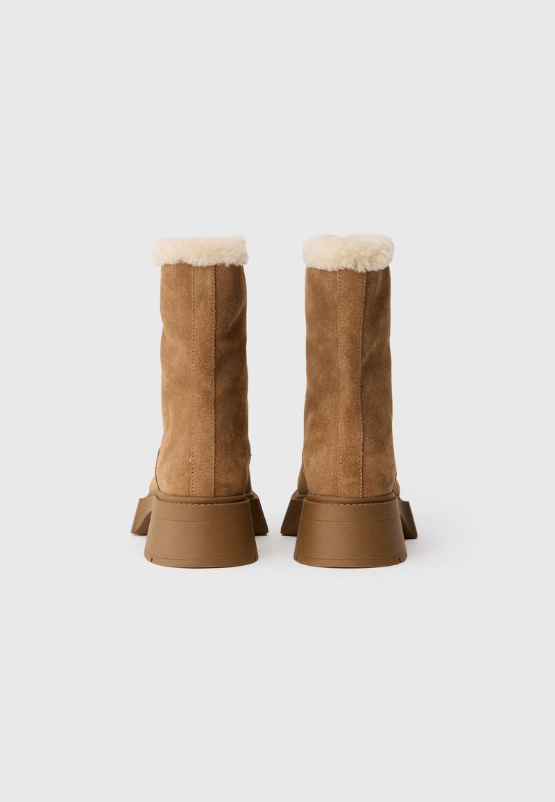 Bruine suède enkelboots met een zachte crèmekleurige voering, een gestructureerde platte hak en een ronde neus. Naaddetails zijn zichtbaar aan de buitenkant.