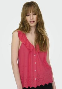 Junge Frau mit langen, welligen Haaren, die eine ärmellose rosa Bluse mit gerafftem Ausschnitt und Lochstickerei trägt und nach unten schaut.