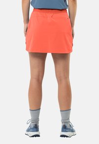 Heldere koraalkleurige rok met een tailleband, voorzien van twee zijzakken. Draagt grijze sokken en blauwe schoenen, wat een sportieve buitenstijl laat zien.