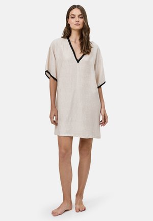 Vrouw staat blootsvoets en draagt een beige jurk tot op de knie met zwarte biezen op korte mouwen en een V-hals tegen een effen achtergrond.